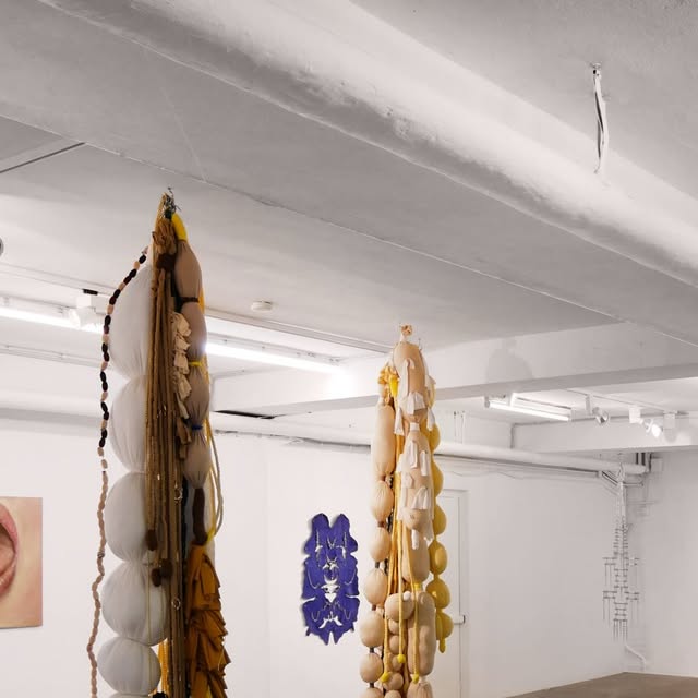 A white room with a low, beamed ceiling and fluorescent tubes. Two long sculptures of stuffed textile forms, beads and cord hang from the ceiling in the foreground. A small painting of a mouth is cut off at the left edge, a blue wall piece hangs on the back wall, and a thin chain-like piece hangs on the right wall.